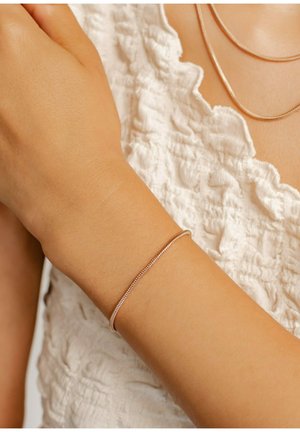 Pulsera de metal color oro rosa con un diseño suave y redondeado, llevada en una muñeca sobre un fondo de tela texturizada blanca. Ajuste relajado.