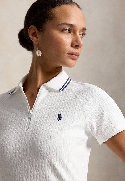 Femeie purtând un tricou polo alb texturat cu un mic logo brodat, privind spre dreapta, cu părul dat pe spate și cercei în formă de scoică.