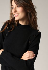 Vrouw met golvend bruin haar, gekleed in een zwarte trui met metallic knopen, die glimlacht en naar links kijkt met gekruiste armen.