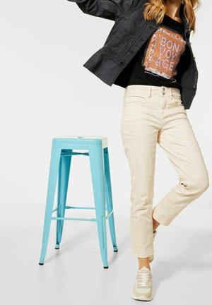 Světle béžové džíny, černé tričko s grafikou "BON VOYAGE" a černá bunda. Vedle oblečení je světle modrá kovová stolička.