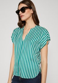 Grønn og hvit vertikalt stripete kortermet bluse med V-hals. Stoffet har en glatt tekstur, kombinert med mørke solbriller.
