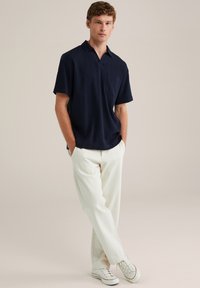 Tricou polo cu mânecă scurtă, de culoare bleumarin, dintr-un material texturat, cu buzunar pe piept și croială lejeră, asortat cu pantaloni albi și adidași.