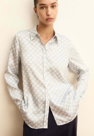 Donna che indossa una camicia bianca con bottoni, decorata con un motivo a catena geometrica azzurro chiaro, in piedi con le mani nelle tasche davanti a uno sfondo uniforme.