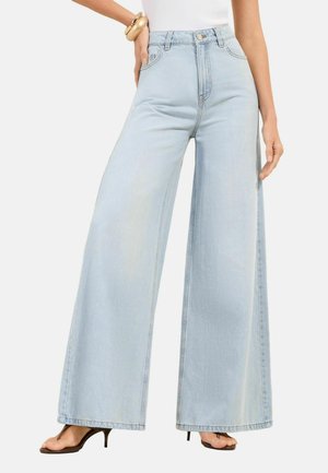 Naine seljas helesinised laia säärtega teksad, valge varrukateta pluus, kuldne käevõru ja tumedate kontsadega sandaalid, seisab ühe käega puusal.