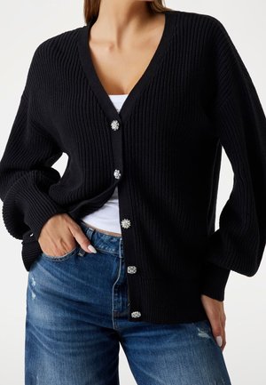 Femme portant un cardigan noir côtelé avec des boutons décoratifs sur un haut blanc et un jean bleu, main dans la poche sur fond clair.