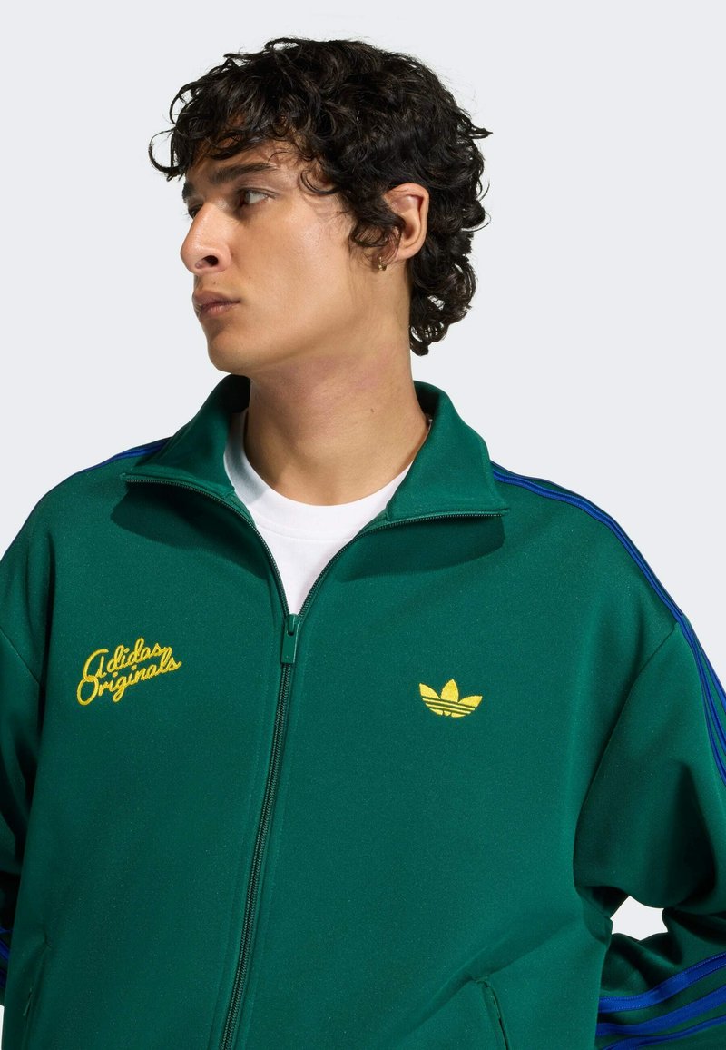Giovane con capelli ricci che indossa una giacca con zip verde Adidas Originals con logo giallo e strisce blu sulle maniche, che guarda verso sinistra.