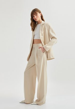 Beige op maat gemaakte blazer over een witte crop top, gecombineerd met losse beige broek. Opvallende details zijn een subtiele zak en stijlvolle schoenen.