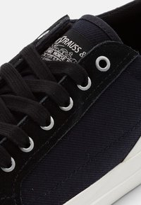 Svart kanvas-sneaker med rund tå, silverfärgade ögon och mörka snören. Har en varumärkesetikett på tungan med vit text och logodetaljer.