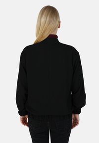 Giubbotto bomber nero con colletto arrotondato, orlo a coste e polsini elasticizzati. Tessuto liscio, dettaglio arricciato in vita, design minimalista.