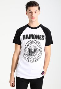 Camiseta de algodón en blanco y negro que presenta un gráfico circular con el texto "RAMONES" y un diseño de águila, mangas cortas y una etiqueta rosa.