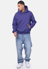 Purple hooded sweatshirt with front pocket, relaxed fit, and ribbed cuffs. Worn with light blue baggy jeans and white sneakers.