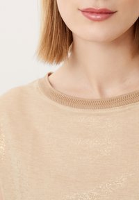 Beige shirt met een soepele textuur, voorzien van een ribbelachtige ronde halslijn. Subtiele glanzende accenten zorgen voor een lichtreflecterende glans op de stof.