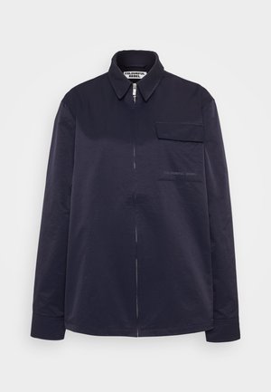 Navy long-sleeve shirt with a zip fastening, pointed collar, and single chest pocket. Features subtle branding on the pocket. Smooth texture.