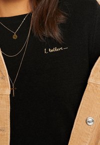 Svart tröja med guld ”jag tror —” broderi, bärs under en ljusbrun kordjacka; lager av silverkedjearmband.