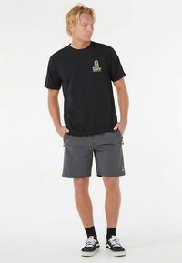 Zwart katoenen T-shirt met een klein gouden logo op de linkerborst, gecombineerd met grijze shorts en zwarte sneakers met witte zolen en sokken.