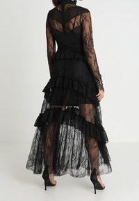 Abito in pizzo nero con maniche lunghe, strati trasparenti, dettagli arricciati e un orlo asimmetrico. Presenta un corpetto aderente e dettagli in pizzo.