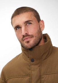 Giacca imbottita marrone con colletto alto, caratterizzata da bottoni neri e dettagli di cucitura. Texture liscia con un design strutturato.