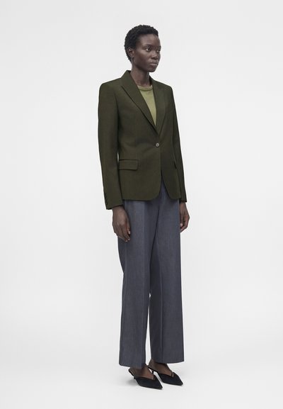 Άτομο στέκεται φορώντας ένα χακί σακάκι, ανοιχτό πράσινο τοπ, γκρι παντελόνι με φαρδιά πόδια και μαύρα μυτερά παπούτσια με λουράκι στο πίσω μέρος σε απλό φόντο.