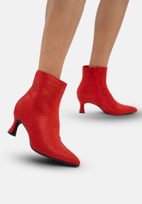 Botines rojos de tobillo hechos de material texturizado con adornos de strass. Punta afilada y un elegante tacón curvado. Cremallera en la parte trasera.