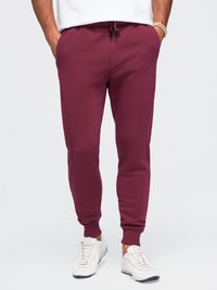 Uomo che indossa pantaloni da jogging bordeaux, sneaker bianche e una camicia bianca, in piedi con le mani in tasca davanti a uno sfondo semplice.