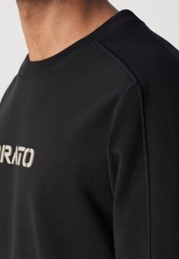Sweat-shirt noir en tissu doux, avec panneaux latéraux côtelés et col rond. Logo "IRATO" affiché en blanc sur le devant.