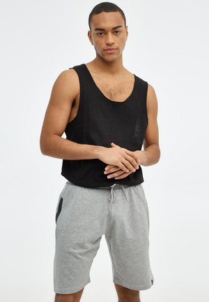 Hombre de pie con las manos entrelazadas, lleva una camiseta negra sin mangas y pantalones cortos grises con cordón, sobre un fondo blanco liso.