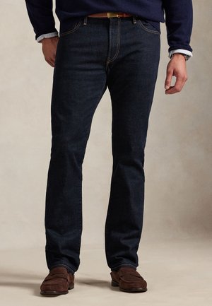 Homme portant un jean bleu foncé, une ceinture marron, un pull bleu marine par-dessus une chemise bleu clair et des mocassins en daim marron, debout devant un fond uni.