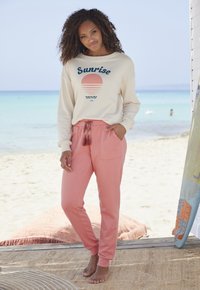 Beige sweatshirt met 'Sunrise' graphic en roze sweatpants met trekkoord en kwastjes. Zacht, casual stof; relaxte pasvorm; twee voorzakken.