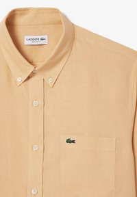 Camicia a maniche corte color arancione chiaro con colletto, realizzata in tessuto testurizzato, con bottoni bianchi e un logo di coccodrillo ricamato verde sulla tasca del petto.