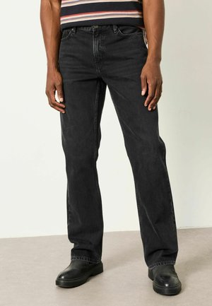 Mann, der schwarze Jeans mit geradem Bein, schwarze Stiefel und ein gestreiftes Hemd trägt, steht auf einem grauen Boden vor einer hellen Wand.