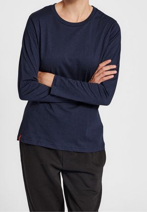 Osoba z założonymi rękami, ubrana w granatową koszulę z długim rękawem i czarne spodnie, na tle jednolitego, jasnego tła.