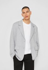Grå, lätt blazer med slagkrage, två framfickor och knappstängning, bärs över en vit t-shirt och svarta byxor.