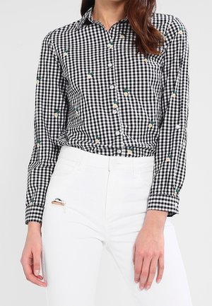 Mujer que lleva una camisa de manga larga a cuadros en blanco y negro con pequeños estampados florales, combinada con pantalones blancos de talle alto con un desgarro cerca del bolsillo.