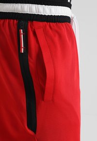 Červené sportovní šortky s černým pasem s bílými a červenými akcenty, kapsou se zipem a texturovanou látkou.