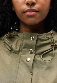 Κοντινό πλάνο του κάτω μέρους προσώπου και του λαιμού ενός ατόμου που φοράει μπουφάν ελιάς με μεταλλικά κουμπιά και ρυθμιζόμενα κορδόνια.