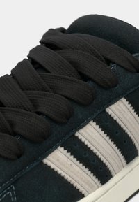 Tmavozelená semišová teniska s čiernymi plochými šnúrkami, vyšivanými bielymi pruhmi a textúrovanou špičkou. Má kontrastnú bielu podrážku.