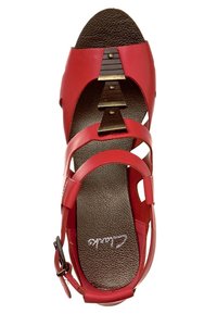 Sandale en cuir rouge avec des brides croisées, semelle intérieure texturée marron et accents métalliques. Elle dispose d'une fermeture à boucle et d'un design ouvert.