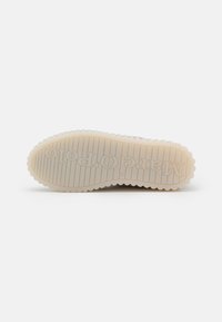 Suela de zapatilla de goma con un patrón estriado texturizado, que presenta una combinación de colores blanco y beige. El nombre de la marca está embosado en la superficie.