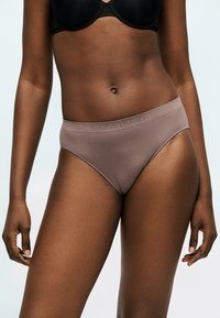 Γυναίκα που φοράει ταυπέ μαγιό υψηλής κοπής Calvin Klein και μαύρο σουτιέν μπροστά σε μονόχρωμο φωτεινό φόντο, δείχνοντας την κοιλιά και τα πάνω μηριαία σημεία.