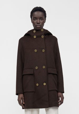 Cappotto doppiopetto marrone scuro con otto bottoni dorati, grandi tasche frontali e cappuccio indossato da una persona in piedi su uno sfondo semplice.