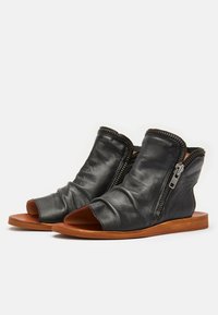 Botas de cuero negro abiertas en la parte delantera con textura arrugada, cremalleras laterales y suelas planas marrones. Presentan un diseño minimalista y una forma única.