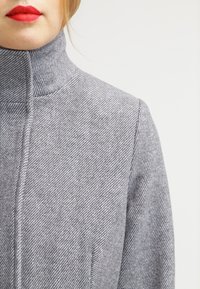 Šedý kabát s vysokým límcem a vzorem rybí kosti, vyrobený z texturované vlněné látky, s detaily švů a bočními kapsami.