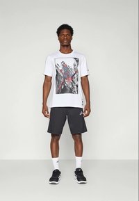 T-shirt branca de manga curta com um gráfico do Spider-Man, combinada com calções pretos. O modelo usa ténis pretos e meias brancas de cano curto.