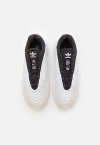 Zapatillas deportivas en blanco y gris con parte superior texturizada, acentos negros, cierre de cordones y una puntera gris clara. Logo de la marca en la lengüeta.