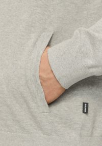 Grauer Sweatshirt mit strukturiertem Finish, ausgestattet mit einer Kängurutasche und gerippten Bündchen. Enthält ein kleines schwarzes Markenschild.