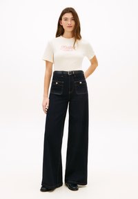 Παντελόνια τζιν με ευρύ παντελόνι σε σκούρο ντένιμ, ψηλή μέση, δύο μπροστινές τσέπες και κουμπιά, συνδυασμένα με εφαρμοστή κρεμ μπλούζα με λογότυπο.