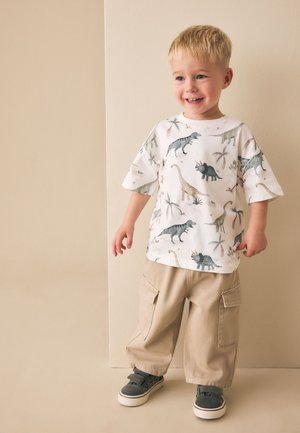 Jongentje lacht, draagt een wit t-shirt met dinosaurusprint, beige cargobroek en grijze sneakers, staat tegen een neutrale muur.