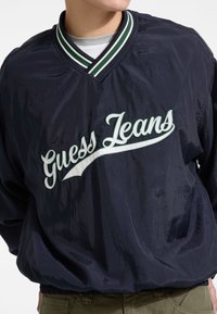 Jūrinis vėjo akiniai su žaliai ir balta dryžuota apykakle, ant krūtinės su dideliu baltu siuvinėtu „Guess Jeans“ tekstu ir elastingomis rankovėmis.