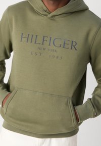 Felpa verde oliva con tasca frontale, polsi e orlo a coste. Presenta il logo "HILFIGER NEW YORK EST. 1985" in blu navy. Tessuto morbido.