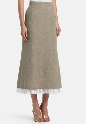 Žena nosi béžovú áčkovú midi sukňu s bielym volánovým lemom, v kombinácii so zlatými sandálmi na vysokom opätku, stojaca na bielom pozadí.